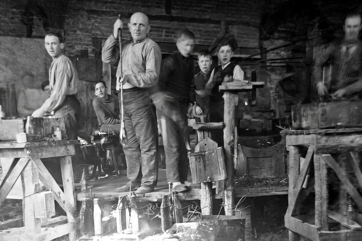 Warsztat huty szkła w Tłuszczu. Butelkę dmucha majster Bolesław Krawczyk, obok stoi pomocnik Stefan Krawczyk, w głębi siedzi Kazimierz Krawczyk.